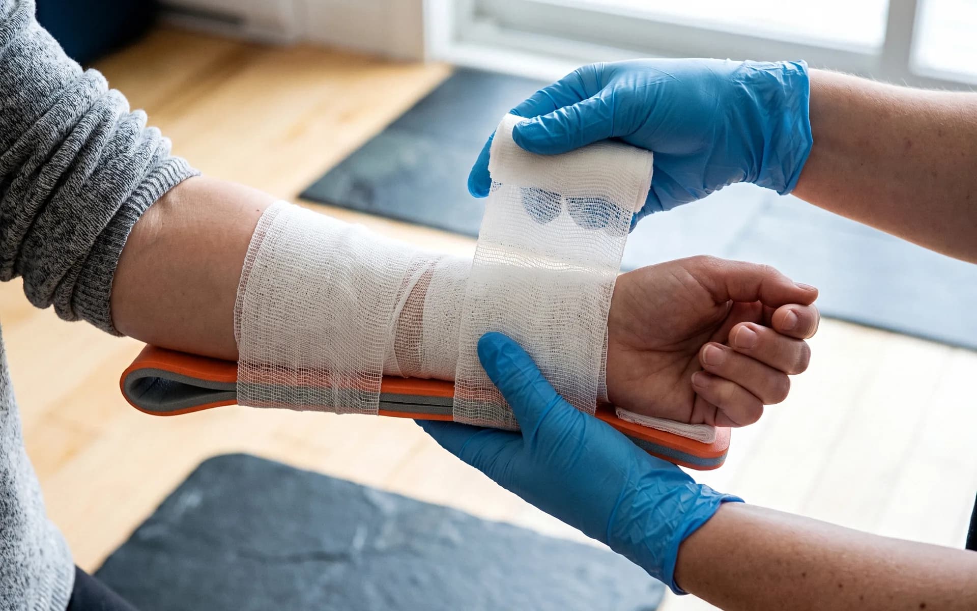 Gloved hands feeding roller gauze over an orange padded splint on a partner's forearm, the partner's hand loosely closed