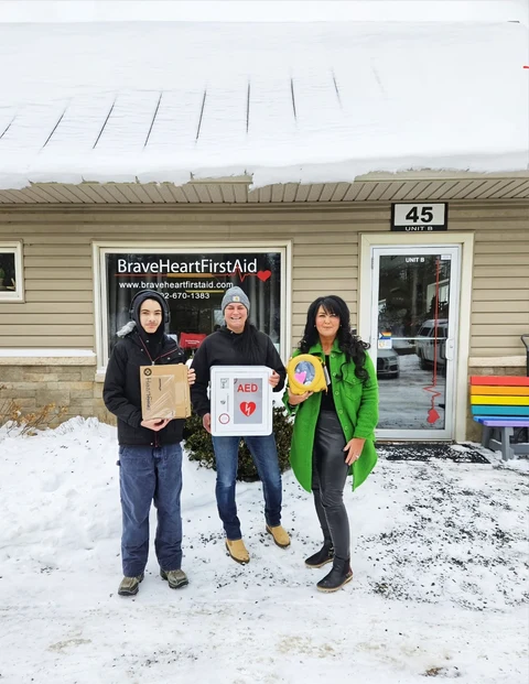 Three people at the Dragonfly Haven AED handover outside BraveHeart, holding the AED, wall cabinet and equipment box.